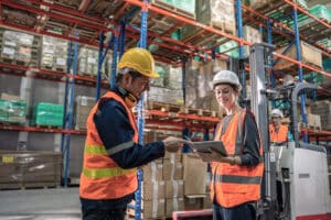 two warehouse workers discussing something on there ipad while a forklift is being operated behind them