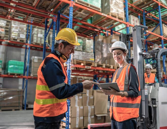 two warehouse workers discussing something on there ipad while a forklift is being operated behind them