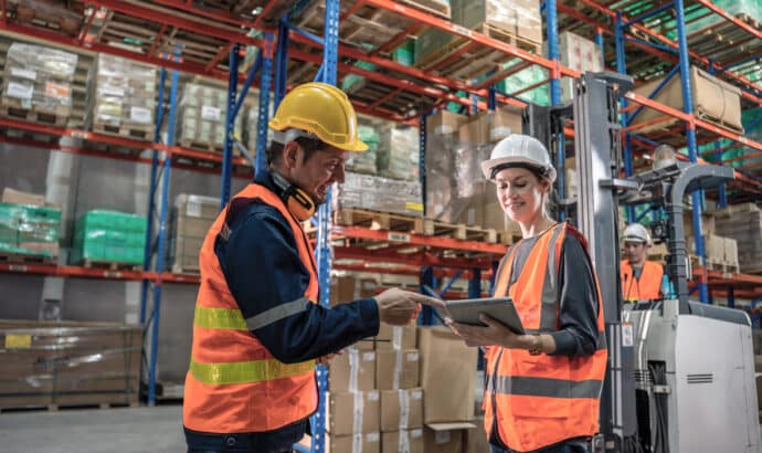 two warehouse workers discussing something on there ipad while a forklift is being operated behind them