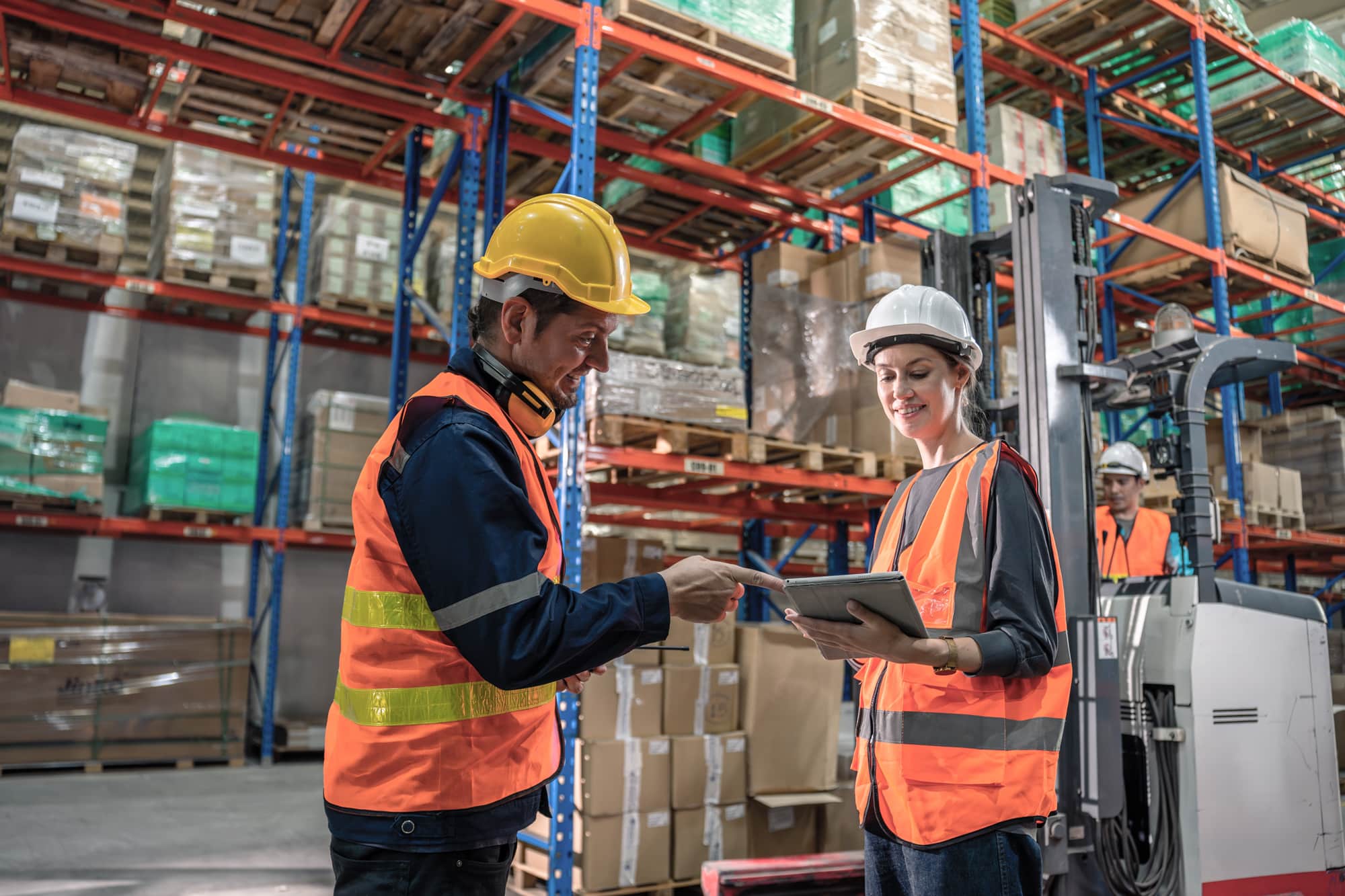 two warehouse workers discussing something on there ipad while a forklift is being operated behind them