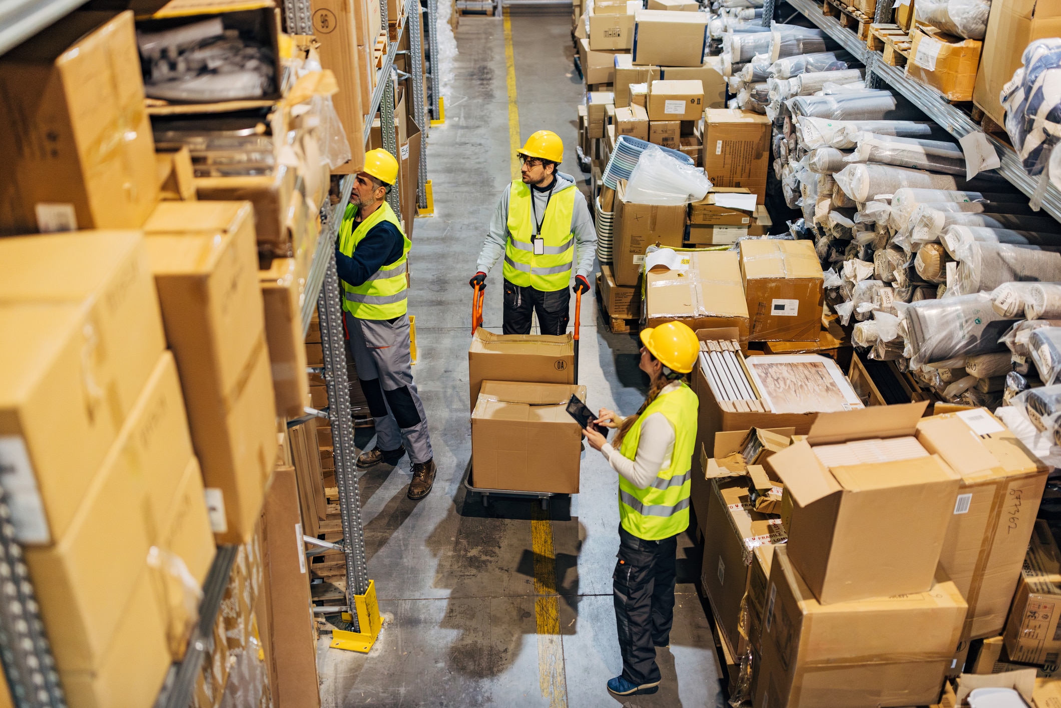 A woman warehouse worker reads a shipment list from a digital tablet while her colleagues prepare the shipment for shipping