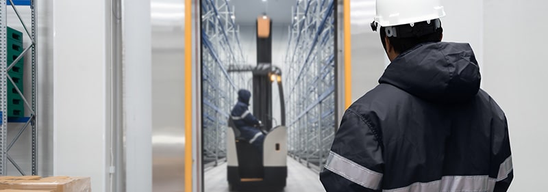 Man in hard hat and coat looking at forklift down the aisle of frozen / cold storage warehouse