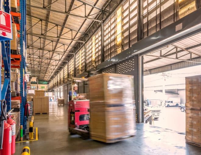 Warehouse worker moving a shrink-wrapped pallet with a forklift near an open loading dock